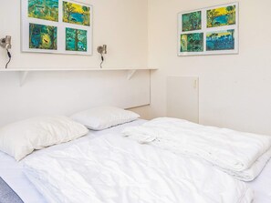 A room with a bed, two pillows, and a wall-mounted shelf with framed pictures.