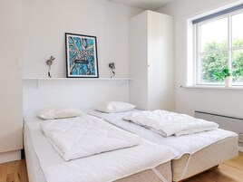 A bedroom with two beds, a window with a view of greenery, a wall-mounted shelf with a framed picture, and a white wall.