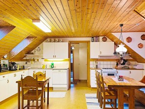 Keittiössä on puupalkit katossa, valkoiset kaapit, ruokapöytä tuoleineen ja riippuvalaisin.