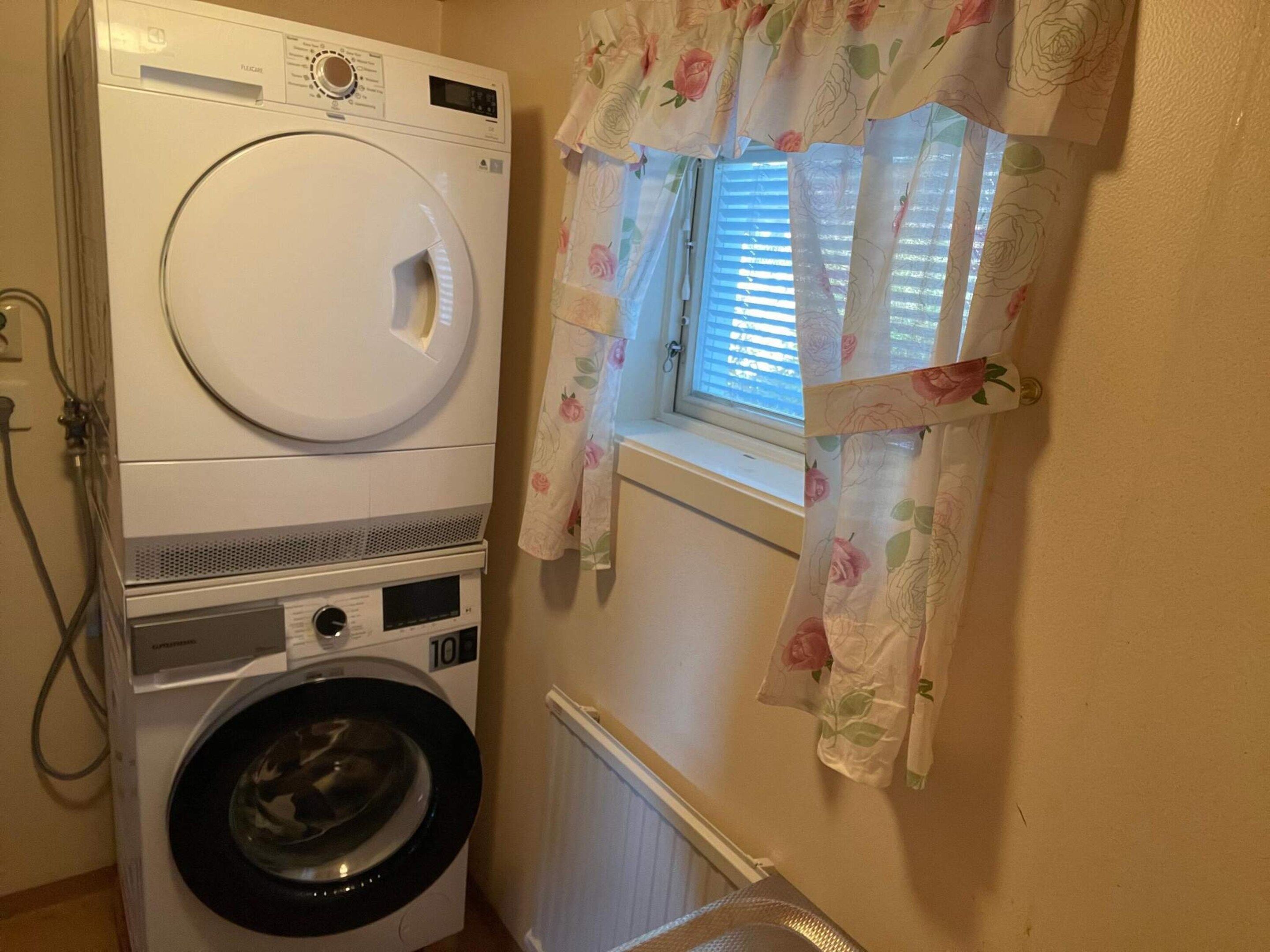 Zona de lavandería con lavadora y secadora apiladas, una ventana con cortinas florales y un radiador.