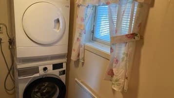 Zona de lavandería con lavadora y secadora apiladas, una ventana con cortinas florales y un radiador.