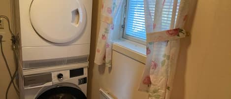 A laundry area with a stacked washer and dryer, a window with floral curtains, and a radiator.