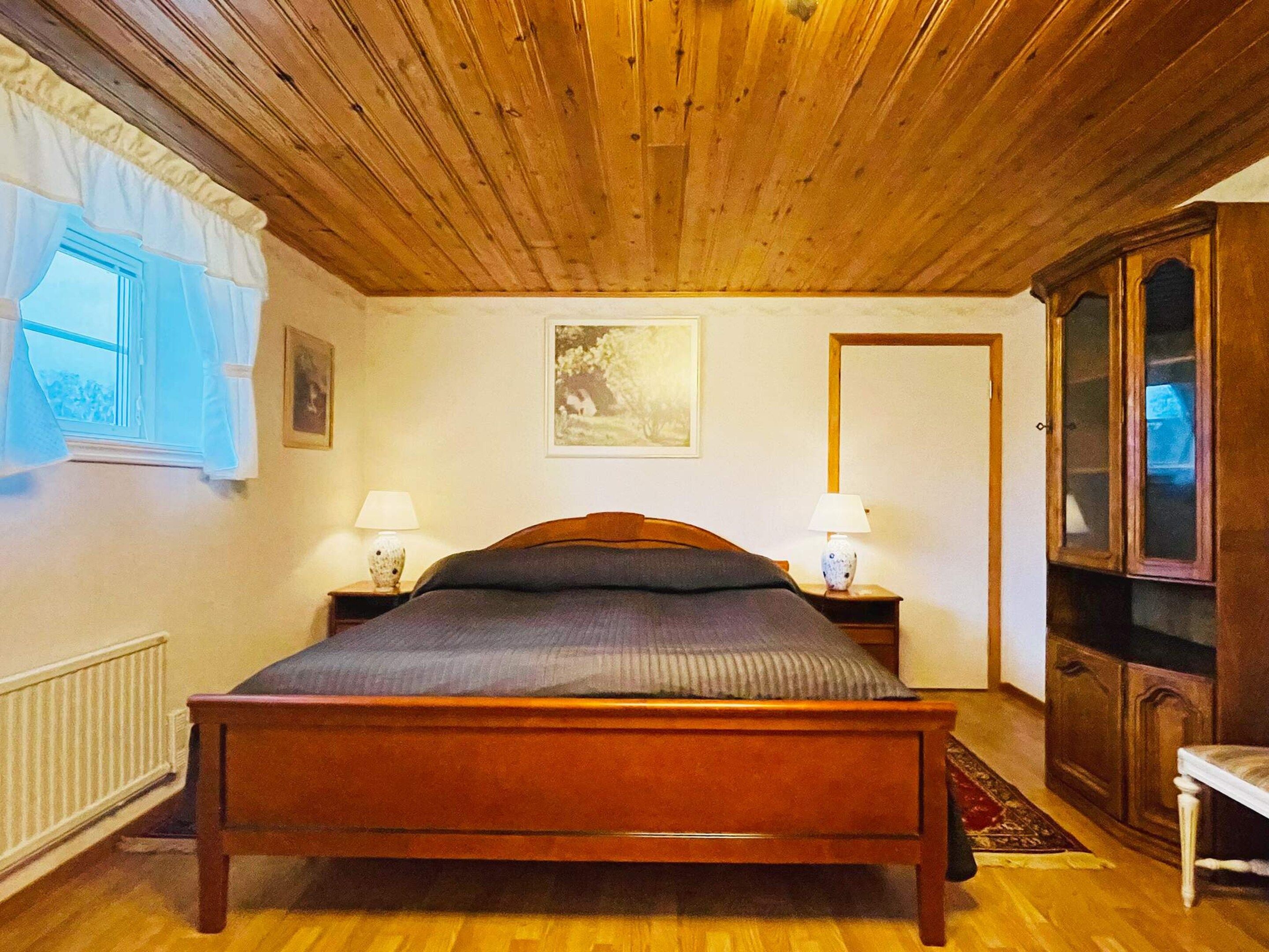 A wooden-paneled bedroom with a bed, two bedside tables with lamps, a radiator, and a cabinet.