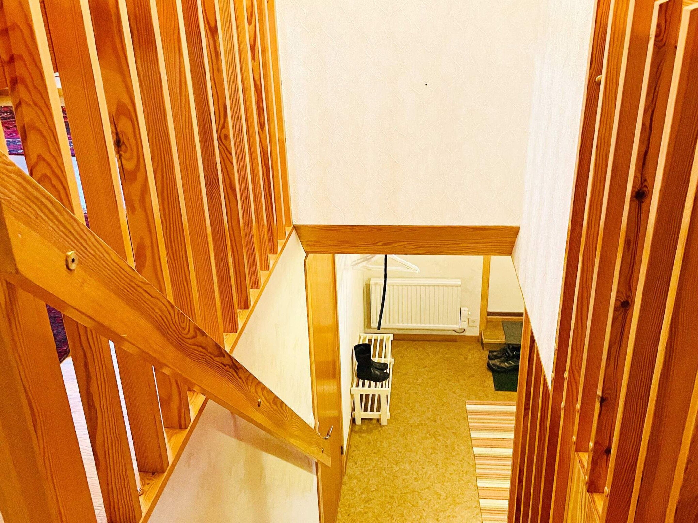 Un escalier en bois mène à une pièce où se trouvent un radiateur et une chaise blanche.