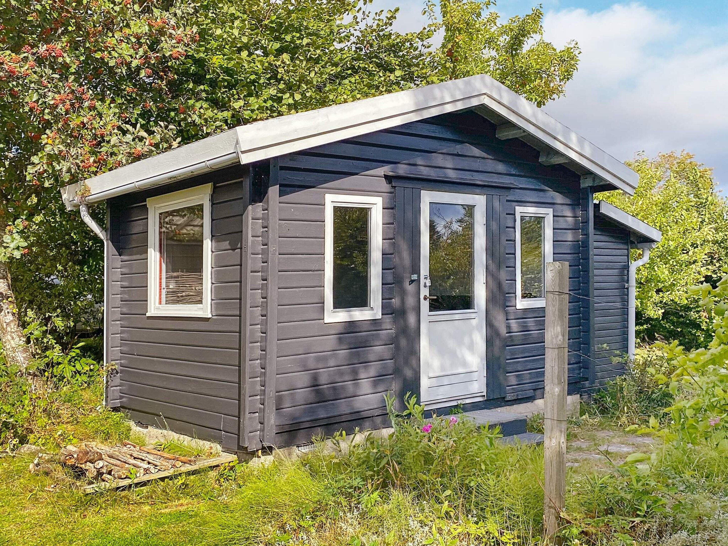 A small, dark-colored cabin with two windows and a door, surrounded by greenery.