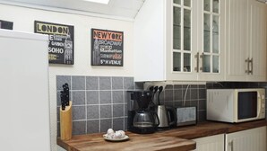 Une cuisine avec des meubles blancs, un four à micro-ondes, une machine à café et un bloc à couteaux. Des panneaux sur le mur indiquent des lieux célèbres, comme Londres et New York.