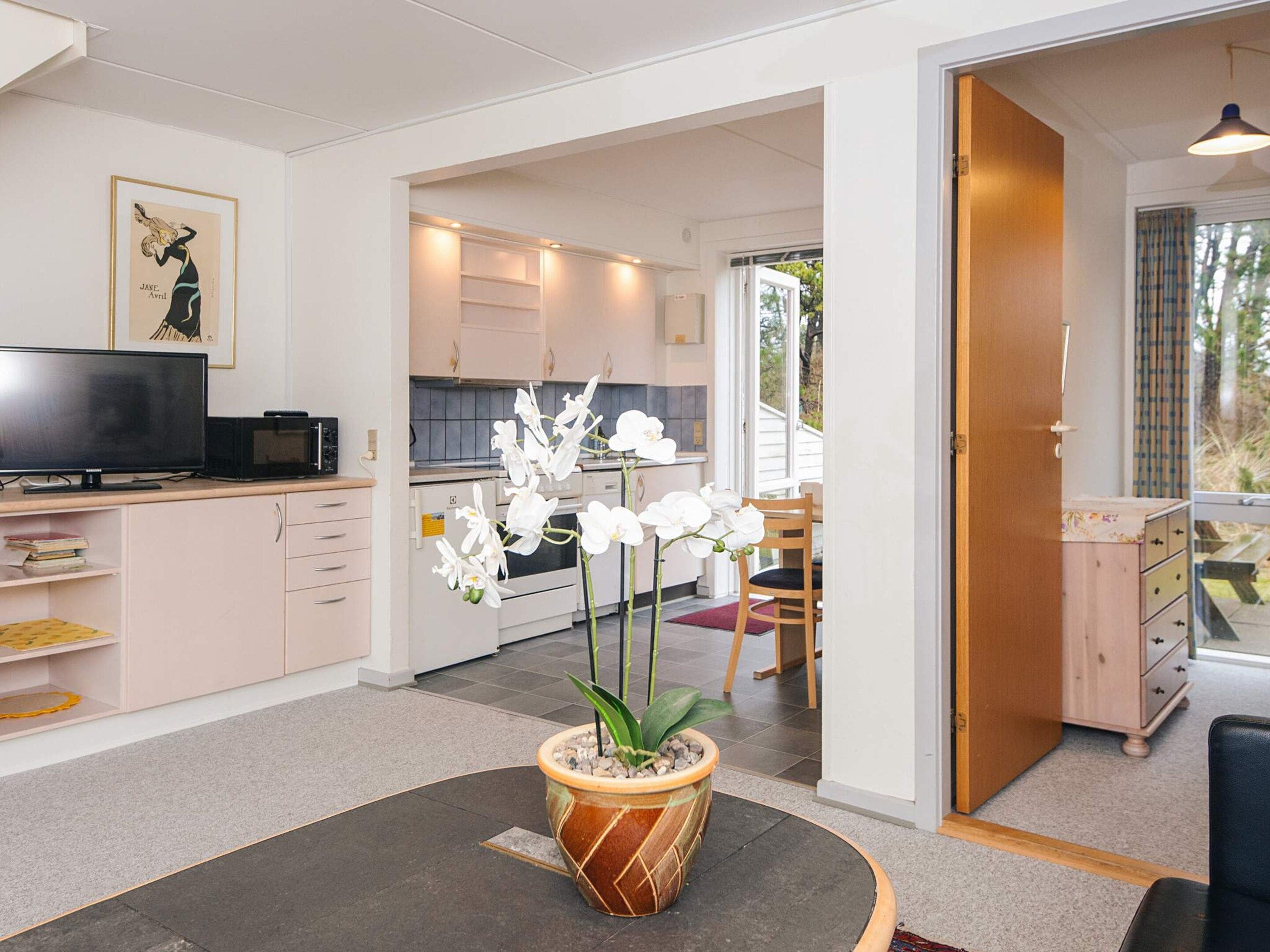 A modern living room with a flat-screen TV, a dining area with a table and chairs, and a kitchen with white cabinets and a microwave.