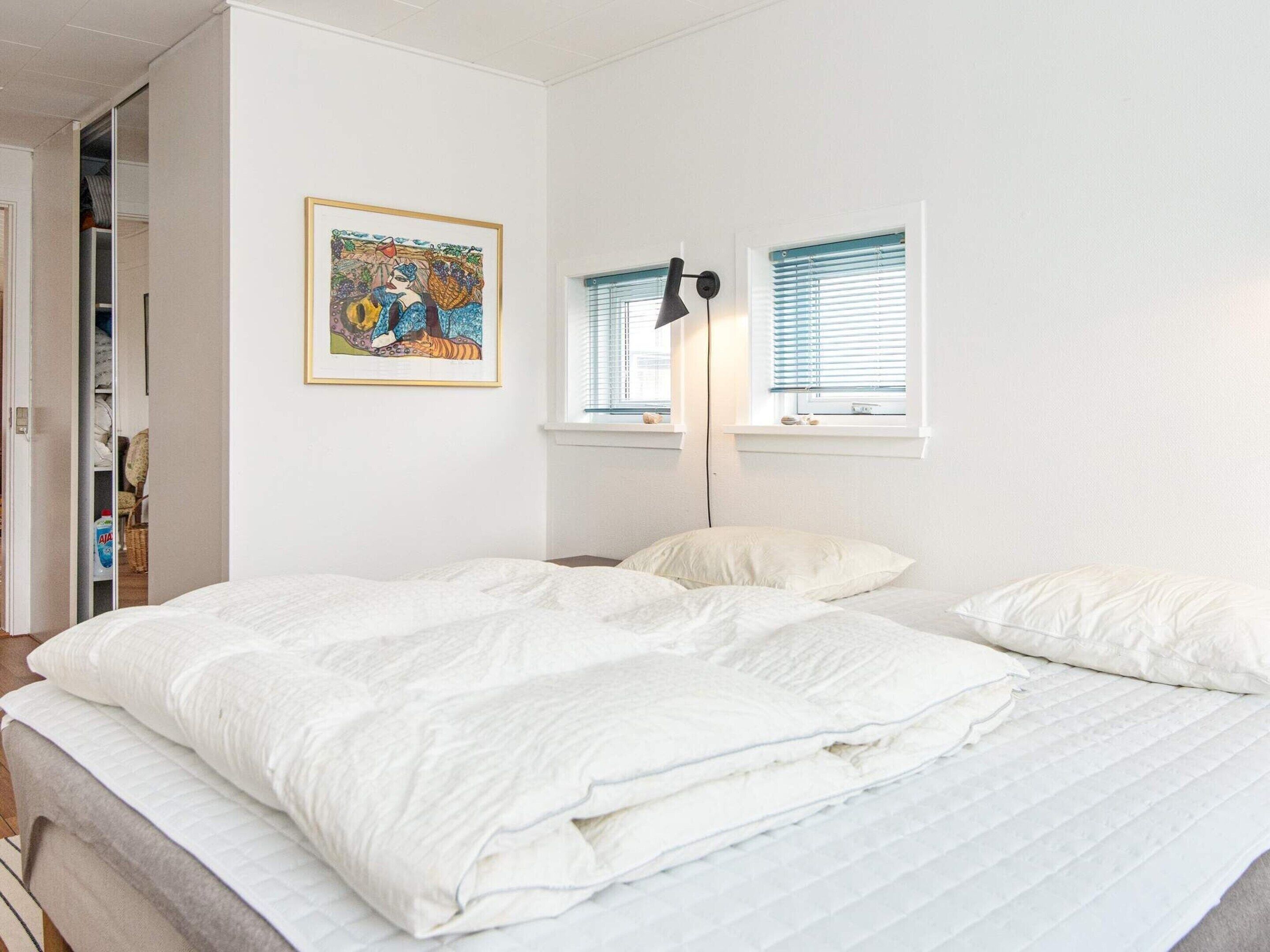 A bedroom with a bed, a nightstand, a lamp, a window with blinds, a door leading to a closet, and a framed picture on the wall.