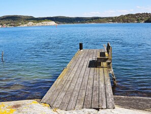 Une jetée en bois s’étendant dans une eau calme, avec une côte rocheuse et des collines en arrière-plan.