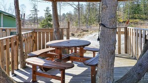 Una terraza de madera al aire libre con una mesa de picnic y bancos, rodeada de árboles y una valla de madera.