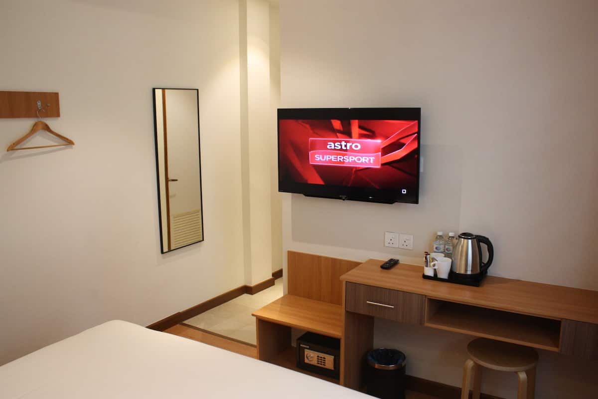 A hotel room with a bed, a wooden desk with a coffee maker, a TV displaying 'astro SUPERSPORT', a mirror, and a hanging coat rack.