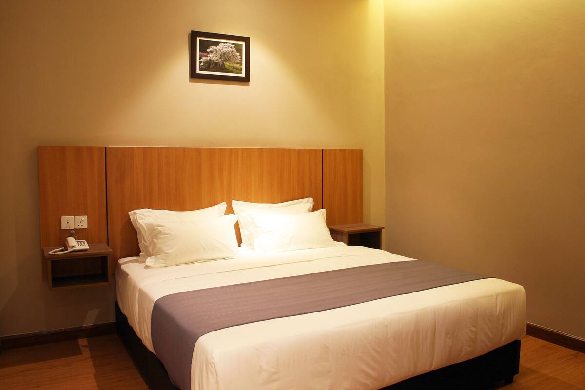 A neatly made bed with white linens and a dark-colored bedspread, a wooden headboard, a bedside table with a phone, and a framed picture on the wall.