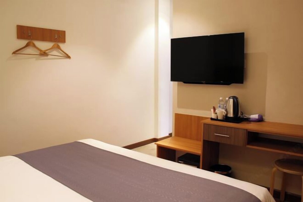 A hotel room with a bed, a desk with a flat-screen TV, a wooden chair, and a coat rack.