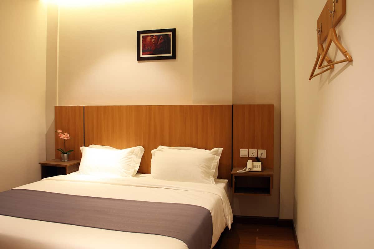 A hotel room with a wooden headboard, a bed with white linens, a bedside table with a phone and a vase of flowers, and a wall-mounted artwork.