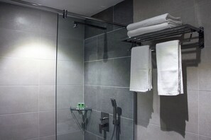 A modern shower area with grey tiles, a glass shower enclosure, and a towel rack with folded towels. - Ahyu Hotel (Kuala Lumpur)