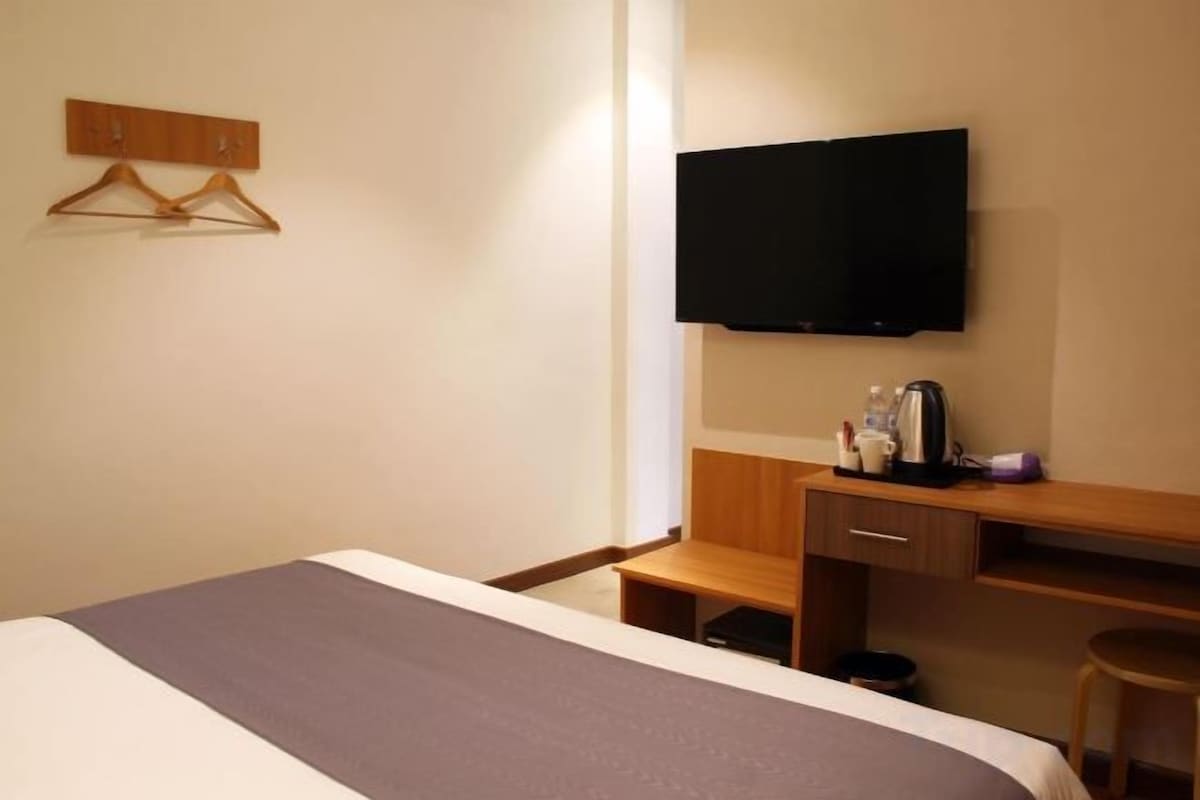 Una habitación de hotel con cama, televisor montado en la pared, escritorio con silla y una mesita.