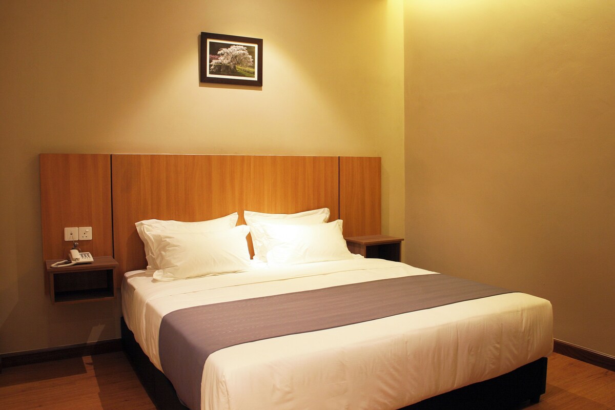 A neatly made bed with white linens and a dark-colored bedspread, a wooden headboard, a bedside table with a phone, and a framed picture on the wall.