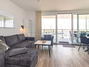 A modern living room with a grey sofa, a small table with a bowl, and a view of a marina through large glass doors. - 5 Star Holiday Home in Wendtorf (Wendtorf)