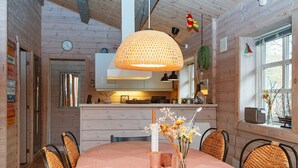 Un coin repas avec une table ronde, des chaises en osier, un luminaire suspendu et une cuisine en arrière-plan.
