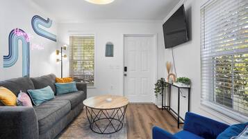 Una sala de estar con un sofá gris, un sillón azul y una mesa de centro redonda. Hay un televisor montado en la pared y una ventana con persianas.