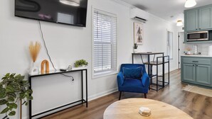 Una sala moderna con televisión, un sillón azul, una mesa de centro de madera y una zona de cocina con armarios color verde azulado y un microondas.