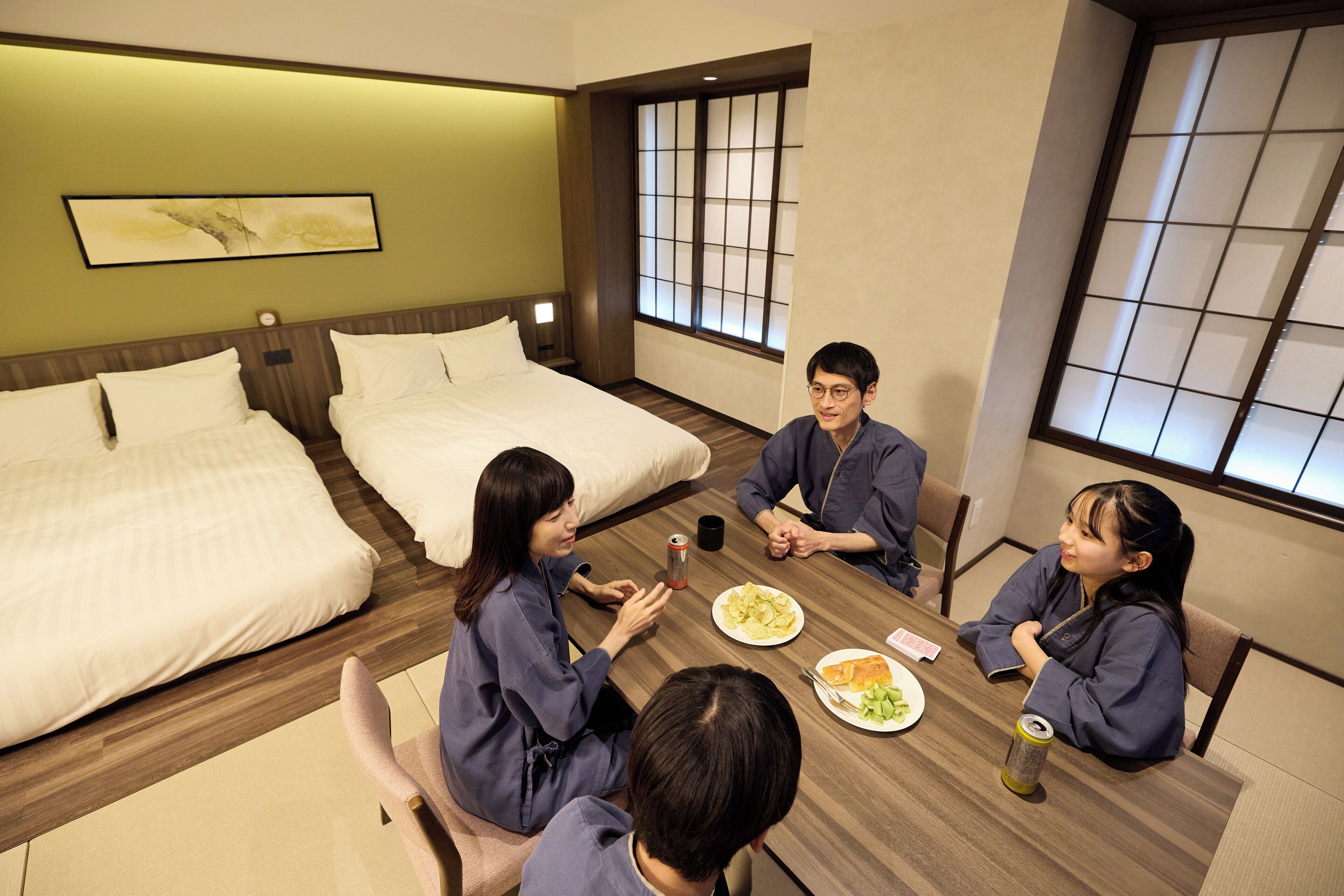 Quarto de hotel com mesa de jantar, quatro pessoas sentadas e uma cama com roupa de cama branca.