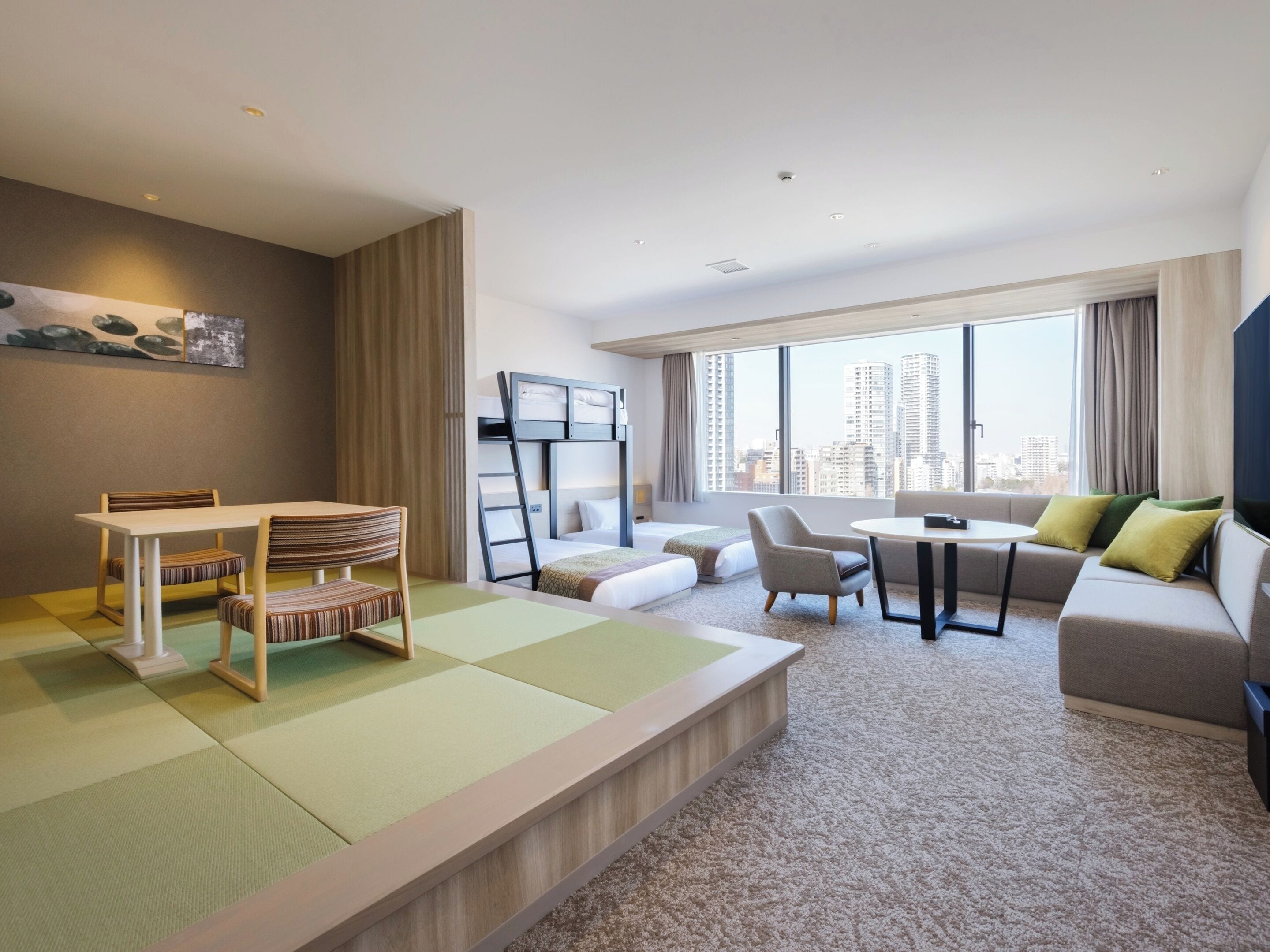 A modern hotel room with a bunk bed, a dining table with chairs, a sofa, and a large window offering a city view.