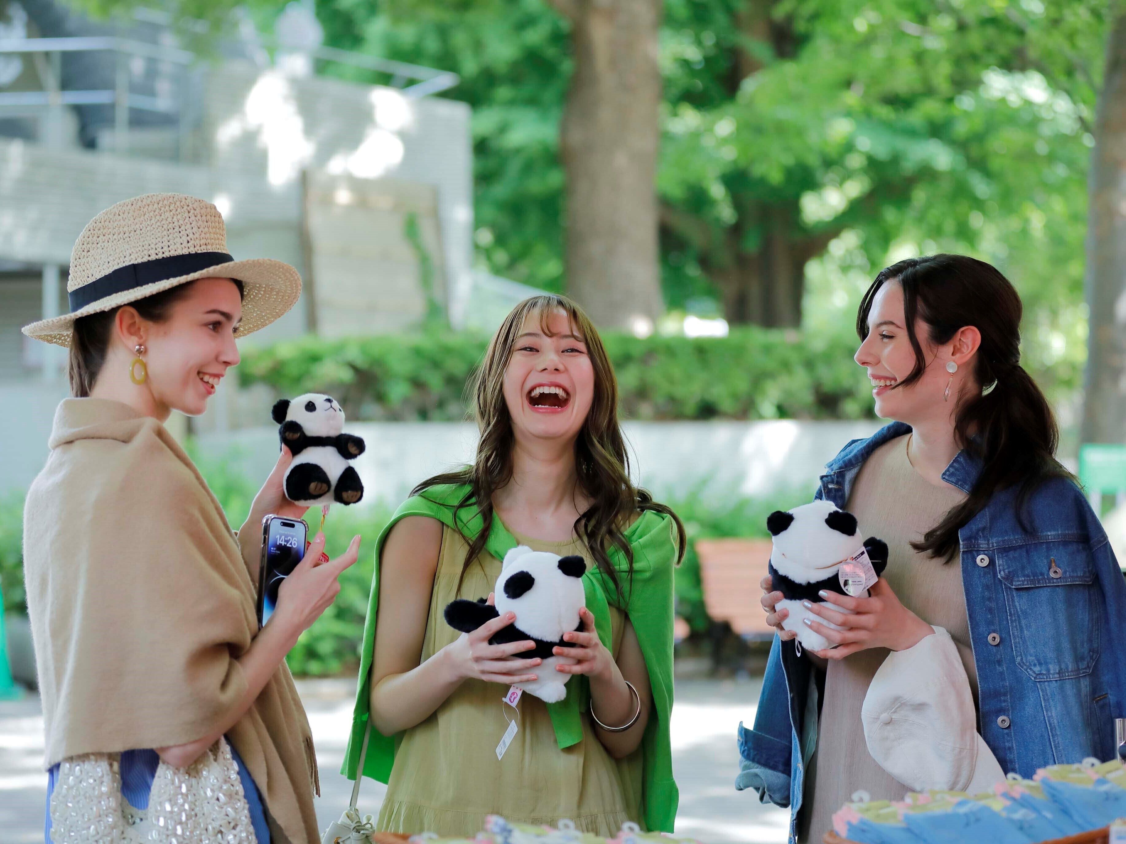 Three people holding panda plush toys in an outdoor setting with greenery in the background.