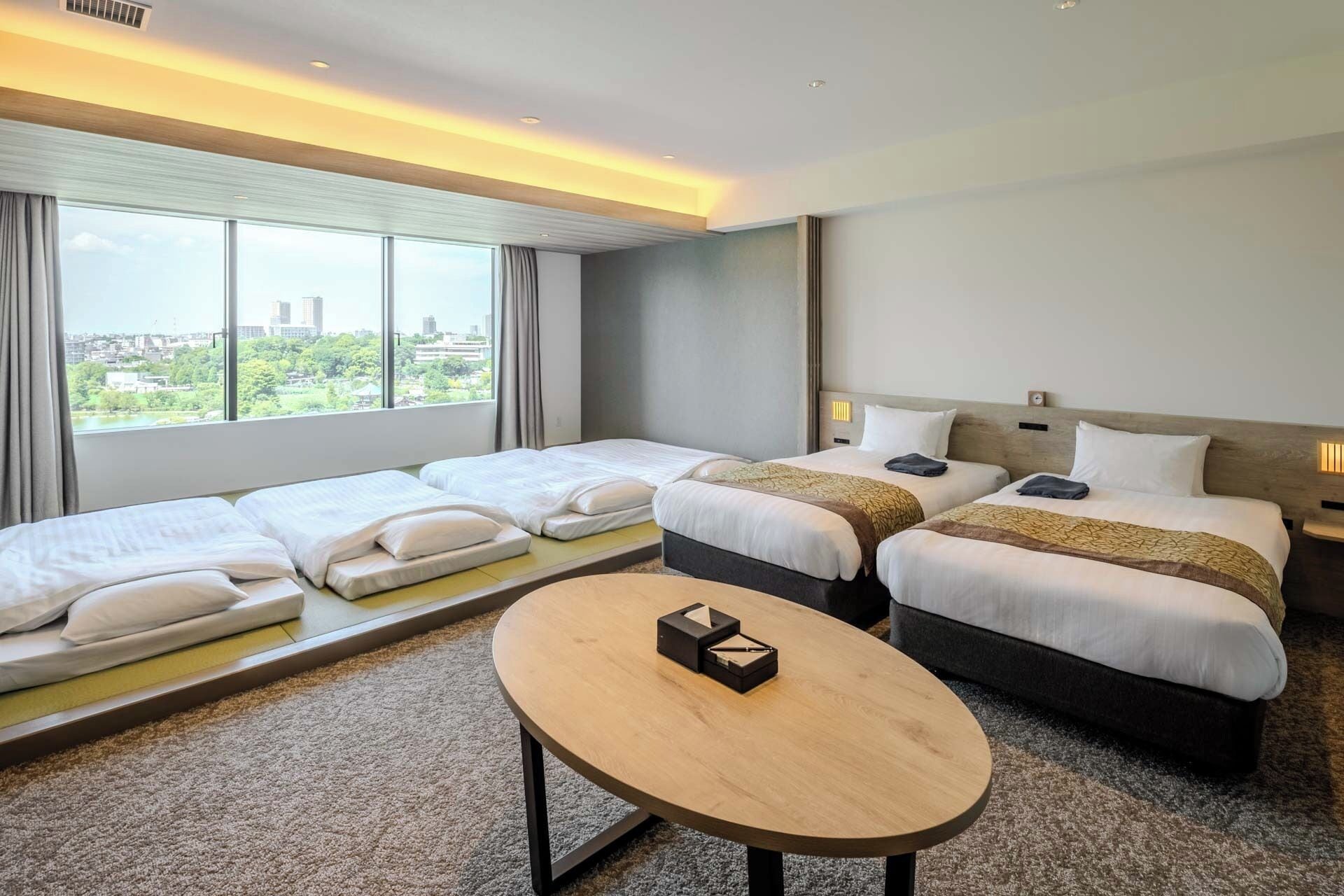 A hotel room with two beds, a small table, and a view of a cityscape through large windows.