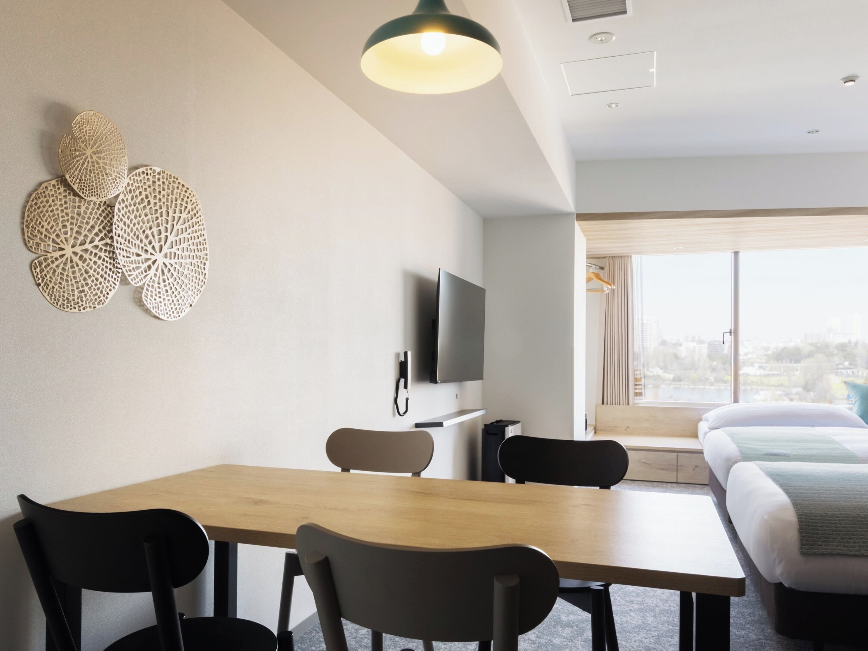 A modern hotel room with a wooden dining table, two black chairs, a flat-screen TV, and a large window with a city view.