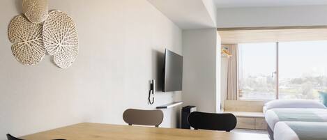 A modern hotel room with a wooden dining table, two black chairs, a flat-screen TV, and a large window with a city view.