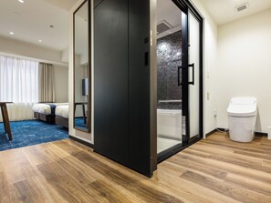 A modern hotel room with a large sliding door to a bathroom, a wooden floor, a bed, a TV, and a trash bin.