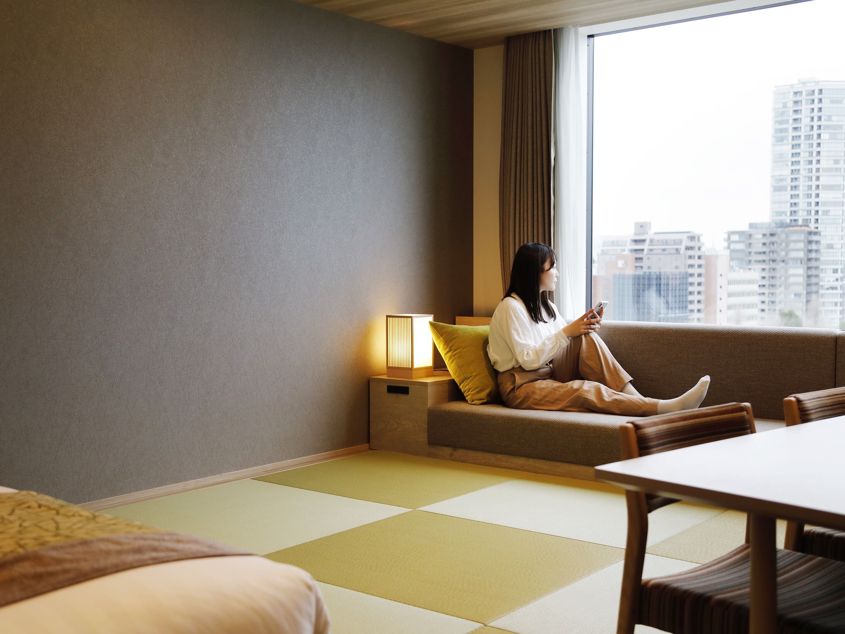 Una mujer sentada en un sofá en una habitación de hotel con vista a la ciudad, una mesa y una lámpara.