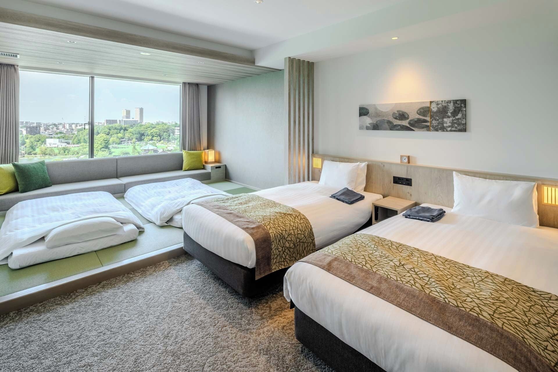 A hotel room with two beds, a large window with a city view, a painting on the wall, and a small table with a lamp.