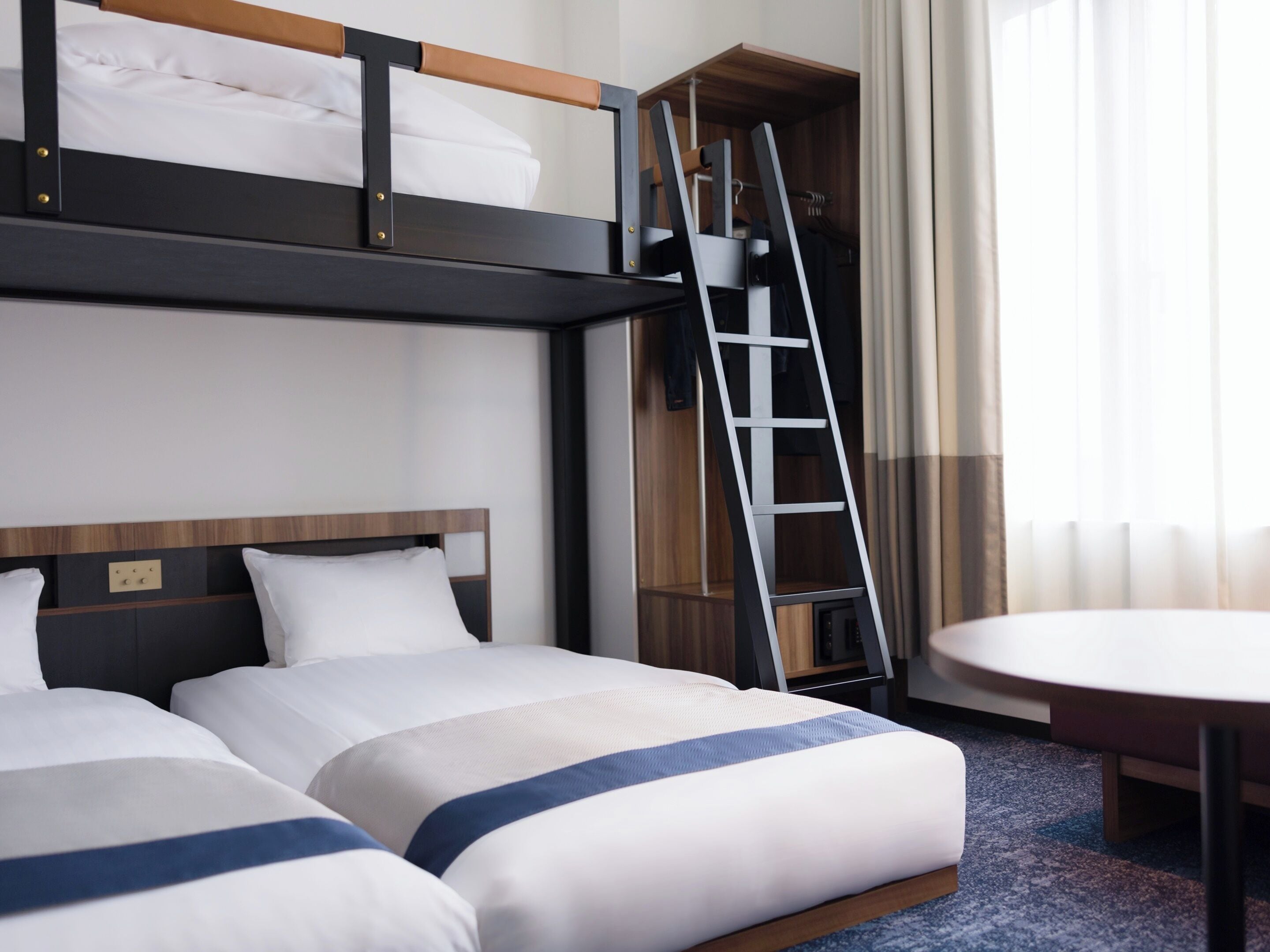 A hotel room with two beds, a wooden headboard, a ladder, a small table, and a window with curtains.