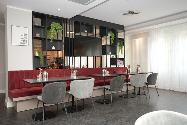 A modern dining area with red upholstered benches, grey chairs, and black tables. There are shelves with plants and decorative items, and a framed quote on the wall. - Boutique Kaiser - Hotel & Apartments (Frankfurt)