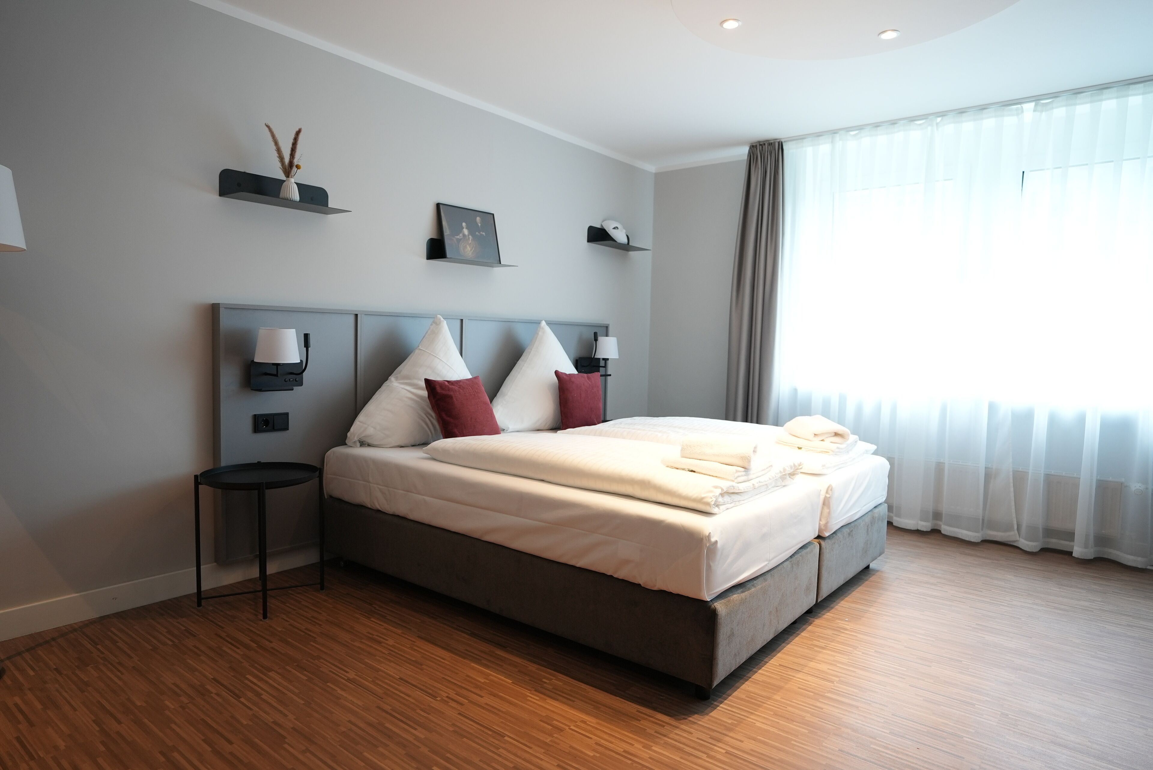 A modern hotel room with a large bed, bedside lamps, a small table, and a window with sheer curtains.