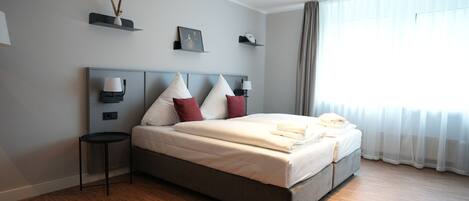 A modern hotel room with a large bed, bedside lamps, a small table, and a window with sheer curtains.