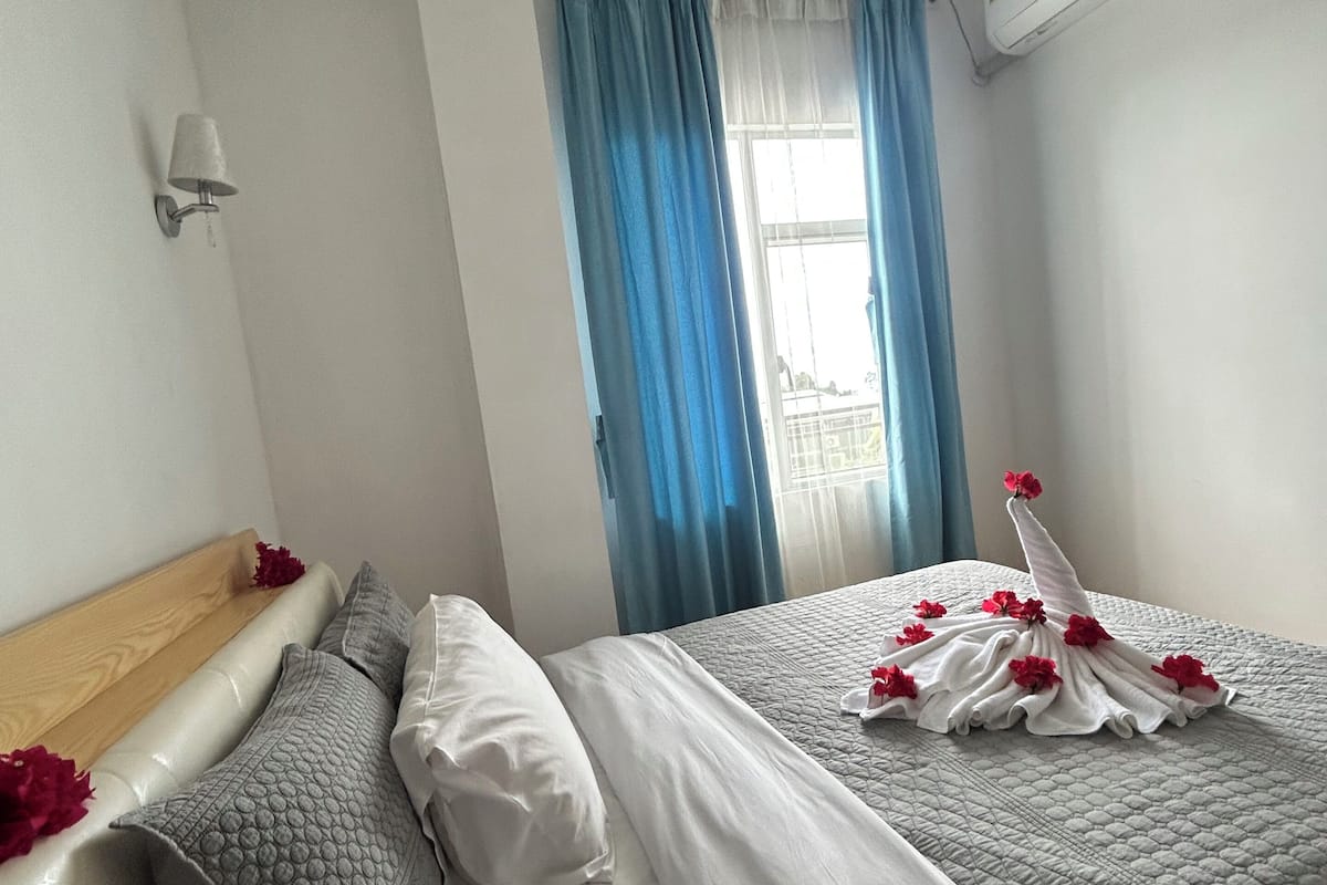Ein Hotelzimmer mit Bett, Kopfteil, einer Wandleuchte, einem Fenster mit blauen Vorhängen und einer Klimaanlage.