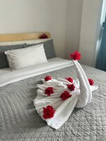 Cama bem arrumada com edredom branco, almofadas cinzas e toalha decorativa em formato de cisne com flores vermelhas.