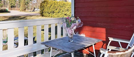 A wooden deck with a table and chairs, a vase with flowers, and a red building in the background.