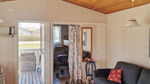 Une pièce avec un plafond en bois, un canapé orné d’un coussin rouge, une table sur laquelle est posé un vase de fleurs, un miroir et une vue sur l’extérieur par une porte ouverte.