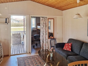 Una habitación con techo de madera, un sofá con un cojín rojo, una mesa con un jarrón de flores, un espejo y vistas al exterior a través de una puerta abierta.