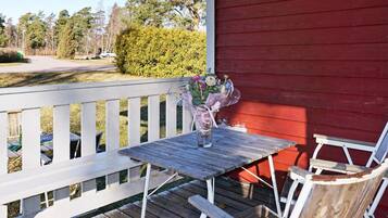 A wooden deck with a table and chairs, a vase with flowers, and a red building in the background.