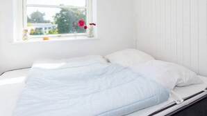 A bed with a white mattress and blue bedding, a window with a view of trees and buildings, and a vase with flowers on the windowsill. - 6 Person Holiday Home in Hjorring (Hjørring)