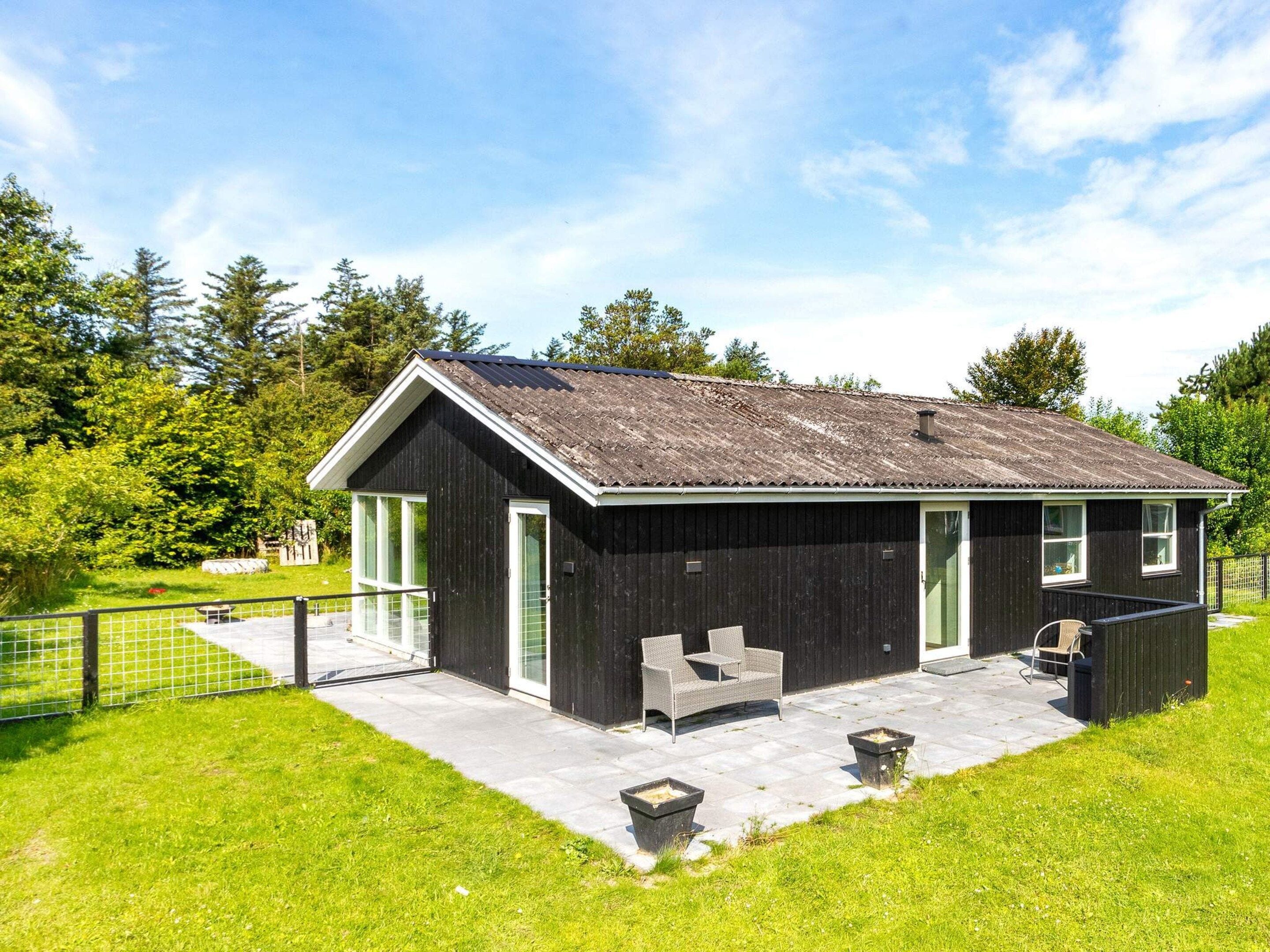 A small, dark wooden building with a sloped roof, a patio with seating, and a fenced garden area.