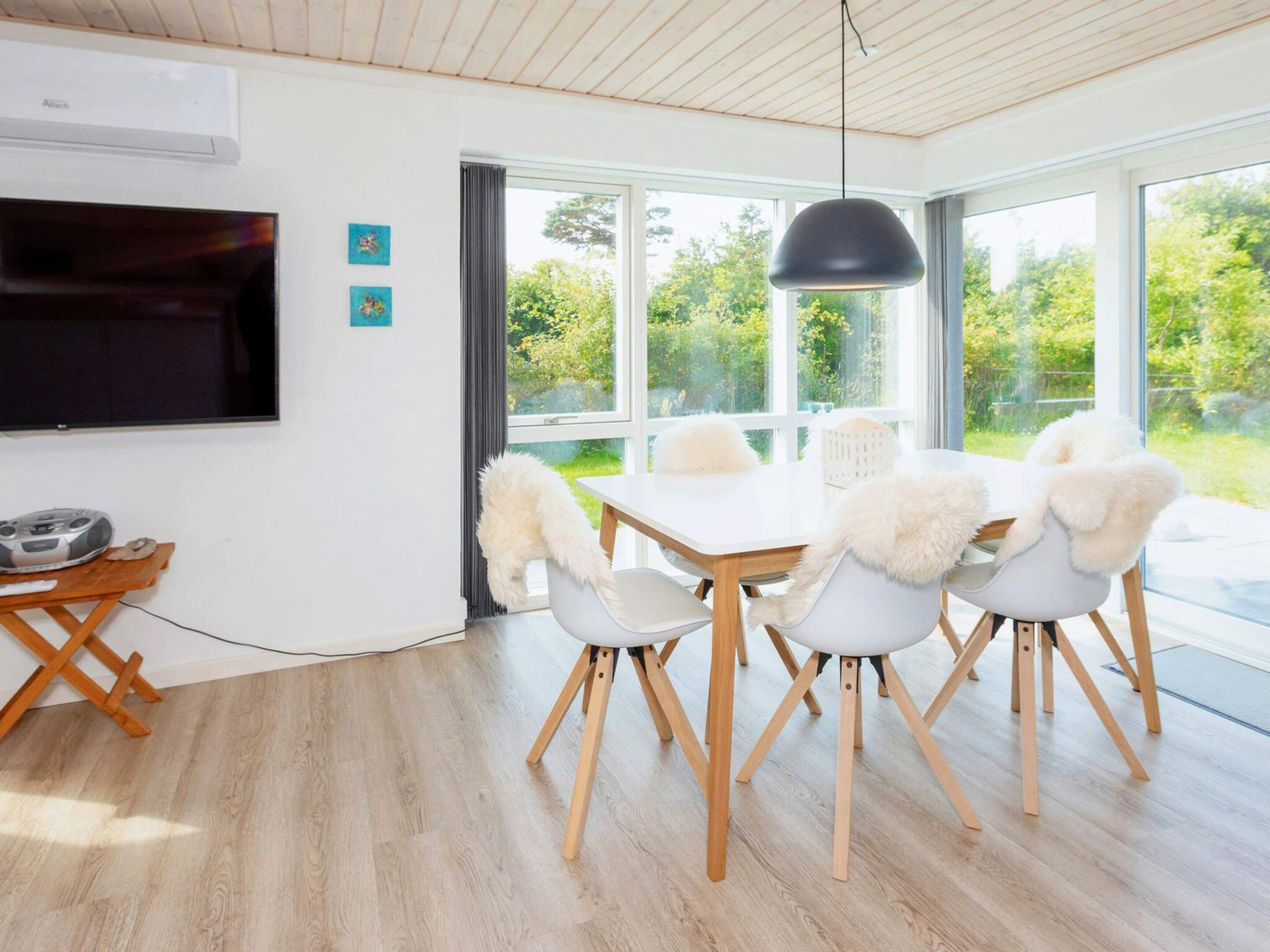 A modern dining area with a white table and chairs, a black pendant light, and a view of greenery outside.