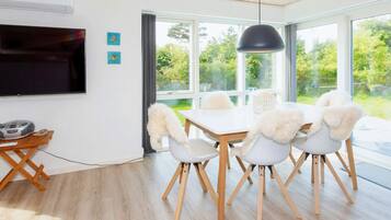 A modern dining area with a white table and chairs, a black pendant light, and a view of greenery outside.