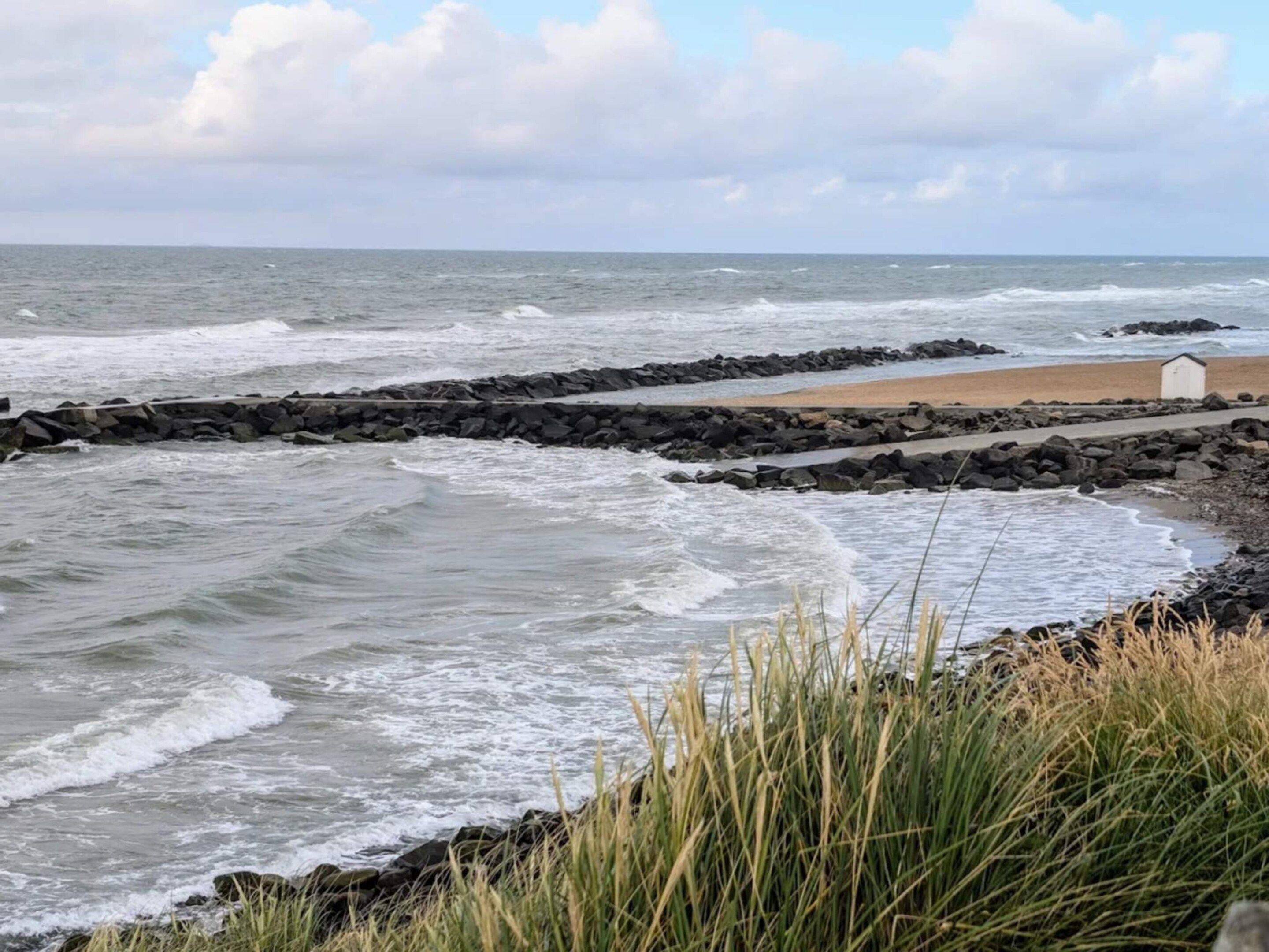 A coastal scene with waves crashing onto a rocky shore, a sandy beach, and a small white building.