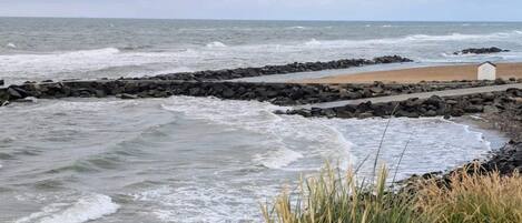 A coastal scene with waves crashing onto a rocky shore, a sandy beach, and a small white building.